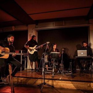 A band performs on a dimly lit stage. A guitarist, bassist, percussionist, and keyboardist are focused on their instruments, creating a relaxed, intimate atmosphere.