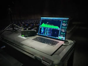 A laptop on a sound technician's table displays audio waveforms with green and blue graphs. Its setting suggests a live music or event venue.