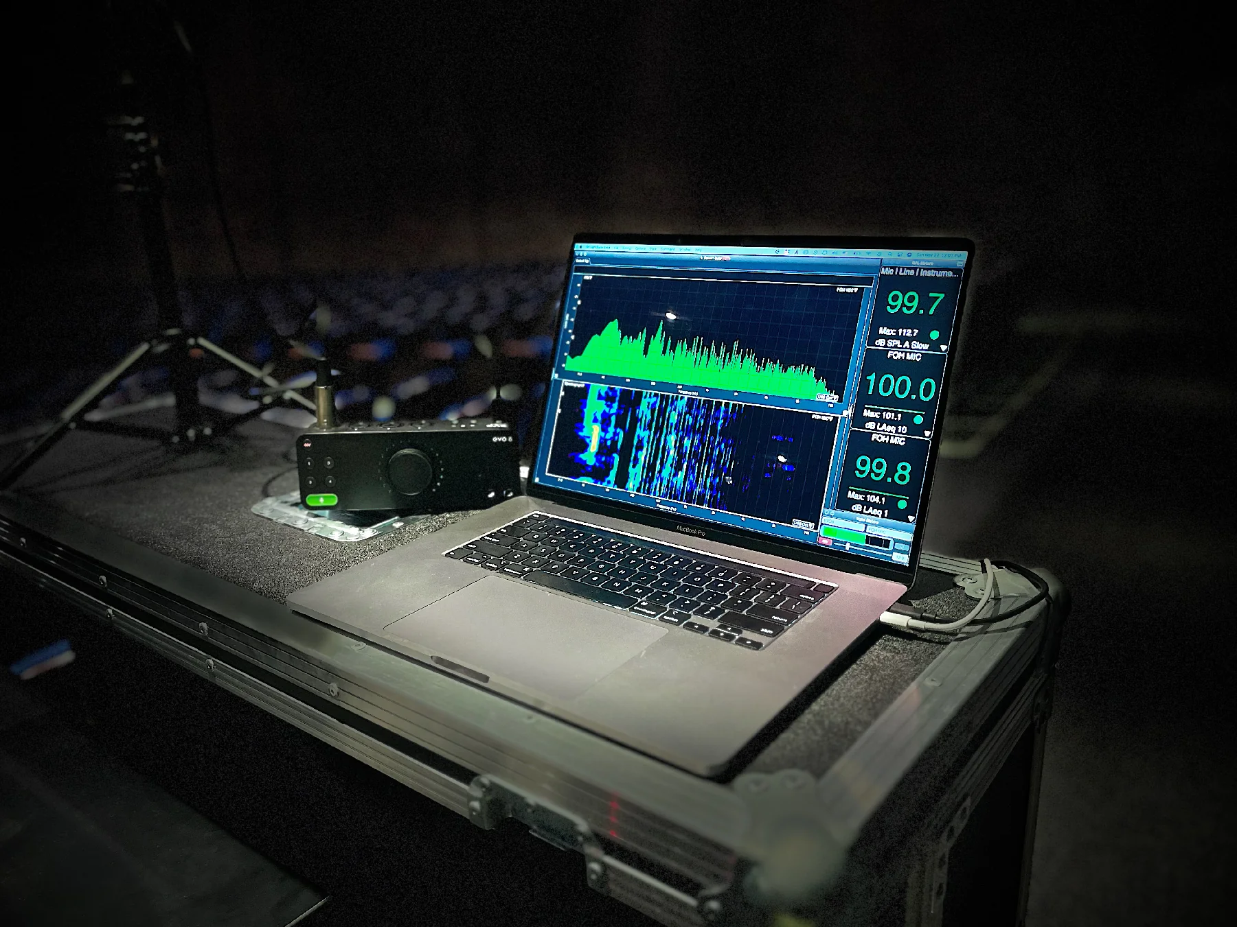 A laptop on a sound technician's table displays audio waveforms with green and blue graphs. Its setting suggests a live music or event venue.